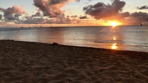 Una playa al atardecer con mar tranquilo, barcos a lo lejos y cielo despejado.