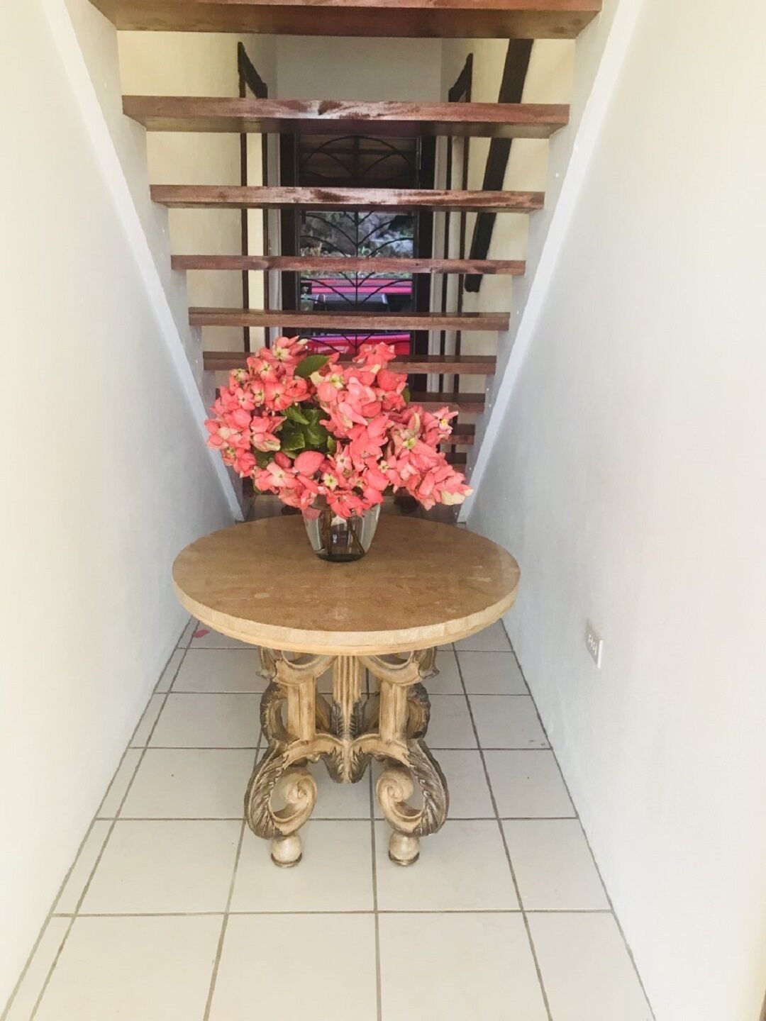 Une petite table ronde surmontée d’un vase de fleurs roses, placée dans un coin, avec un escalier en arrière-plan.
