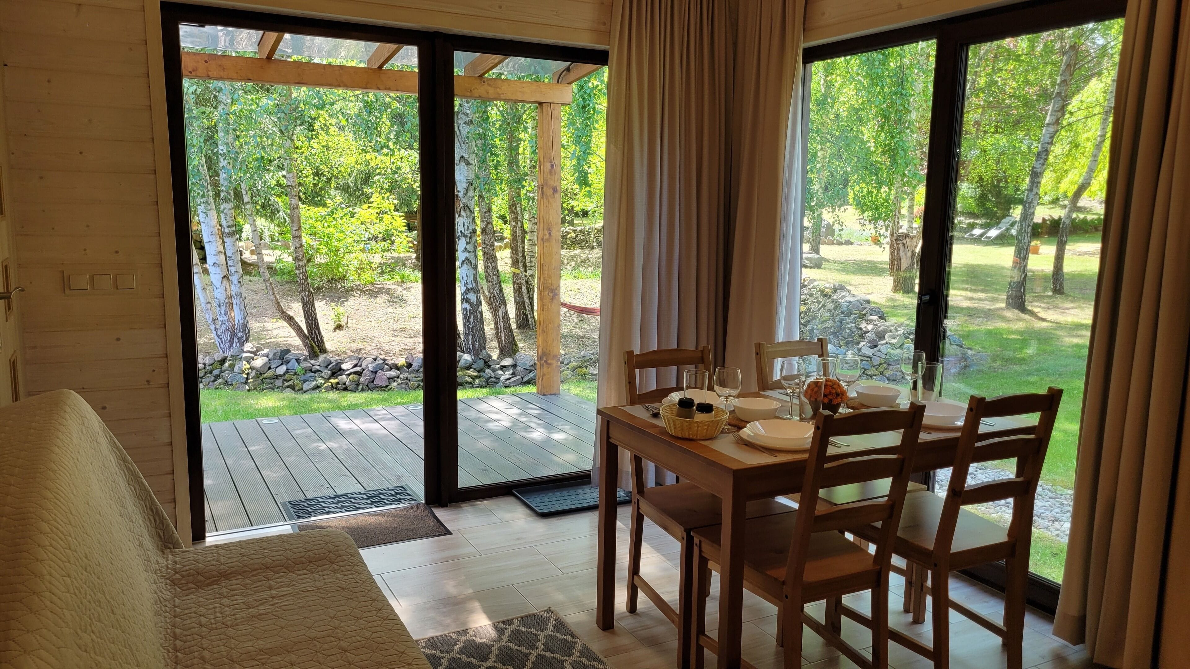 Une pièce avec une table en bois dressée pour le repas, un canapé et une vue sur un jardin grâce à de grandes fenêtres.