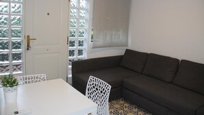 A room with a brown sofa, a white table, and a white chair. There is a door with a window and a view of greenery outside. - D2 Ducal Apartment with private pool (Gandia)