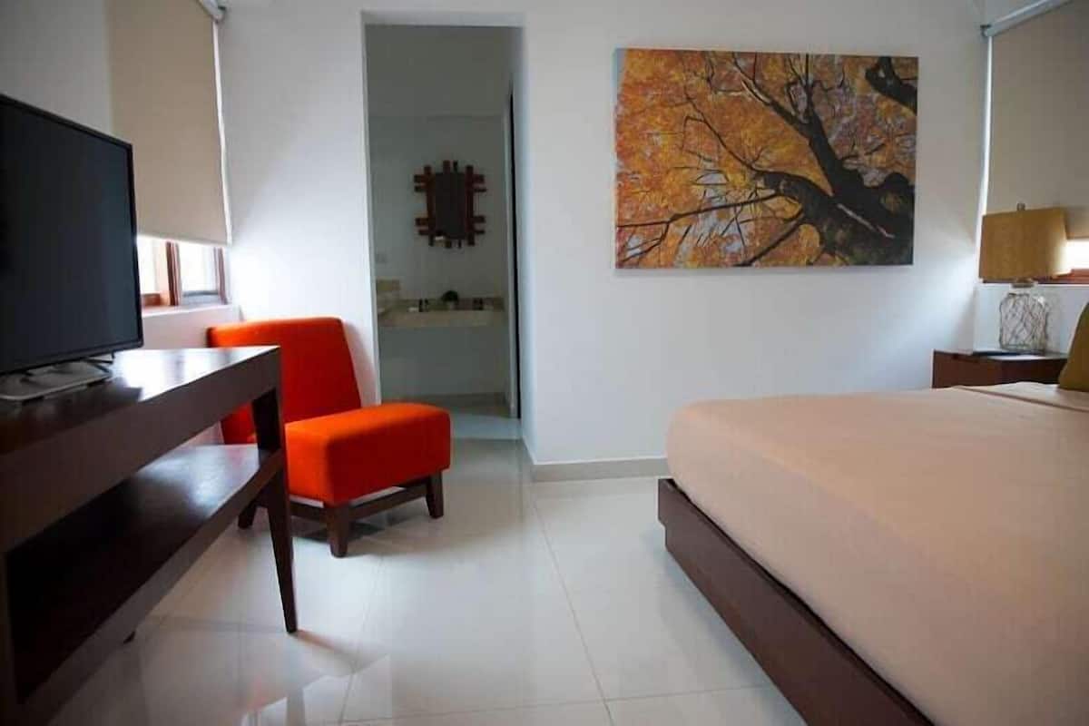 A hotel room with a bed, a desk with a computer, an orange chair, and a painting on the wall.