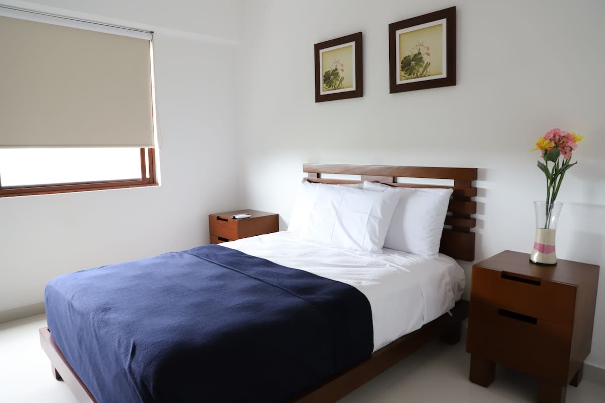 A bedroom with a wooden bed, white bedding, a nightstand, a vase with flowers, and two framed pictures on the wall.