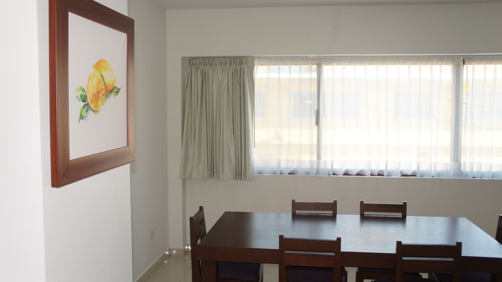 A dining area with a wooden table and chairs, a window with curtains, and a framed picture on the wall.