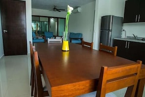 Uma área de refeições com uma mesa de madeira, cadeiras e um vaso com flores. Ao fundo, há uma cozinha com armários e eletrodomésticos.