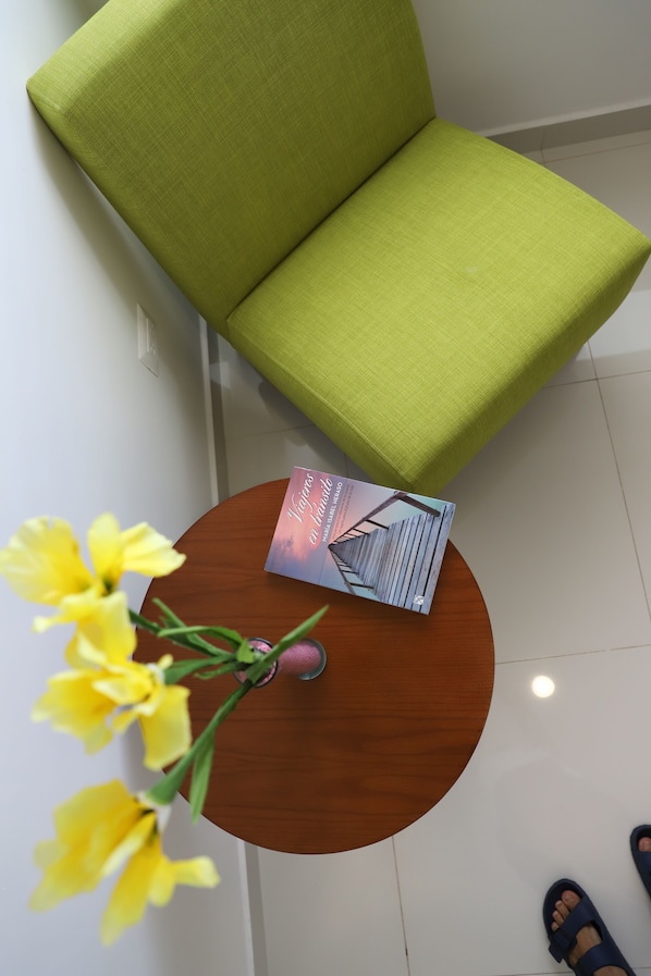 Una silla tapizada de verde, una mesa de madera con un libro y un jarrón de flores amarillas.