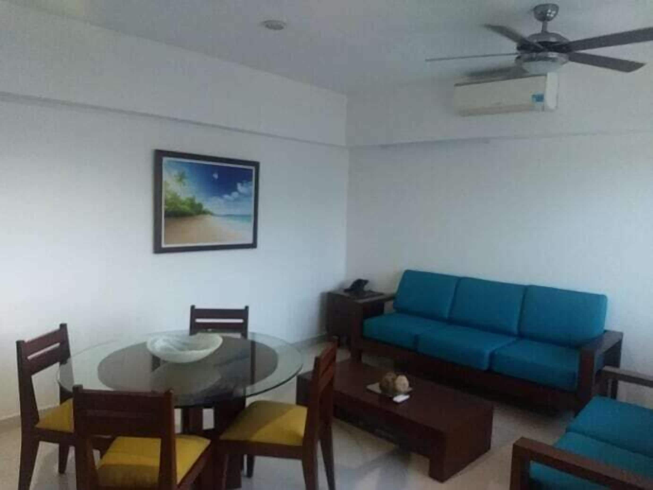 A living room with a glass dining table, blue sofa, and a ceiling fan.
