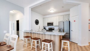 A modern kitchen with white cabinets, stainless steel appliances, and a marble countertop. There are four wooden stools and a dining table with chairs. - Spacious and Lovely 3BD Unit With Balcony, Close to the Convention Center #3vc309 (Orlando)