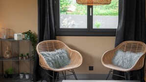Una habitación con dos sillones de mimbre con cojines grises, una mesa blanca, una estantería metálica con plantas y una ventana que da a zonas verdes.