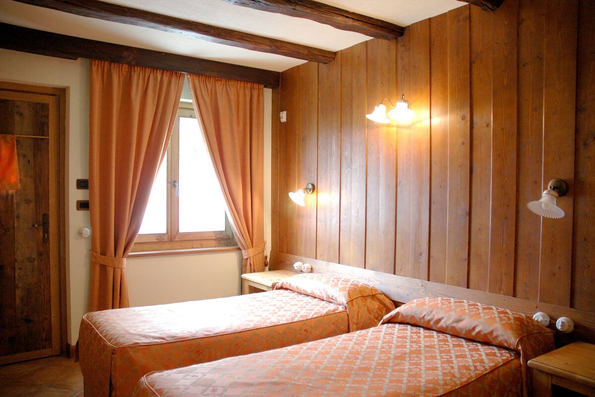 Una habitación con dos camas, paredes de madera, una ventana con cortinas e iluminación en la pared.