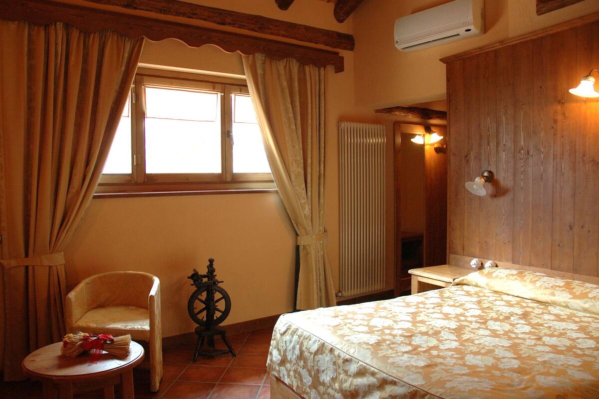 Una habitación con cama, una silla, una mesita con una cesta, un armario de madera y una ventana con cortinas.
