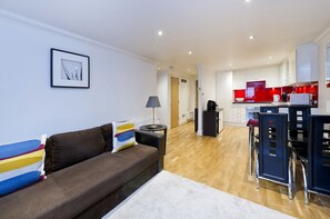 A modern living room with a brown sofa, a small side table, and a framed picture on the wall. A hallway leads to a kitchen with white cabinets and red accents. - 3, Gallery Court · 1 Bed Chelsea Flat + Communal Terrace (London)