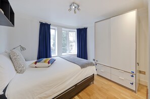 A bedroom with a bed, a bedside table, a wardrobe, and a window with curtains. - 3, Gallery Court · 1 Bed Chelsea Flat + Communal Terrace (London)