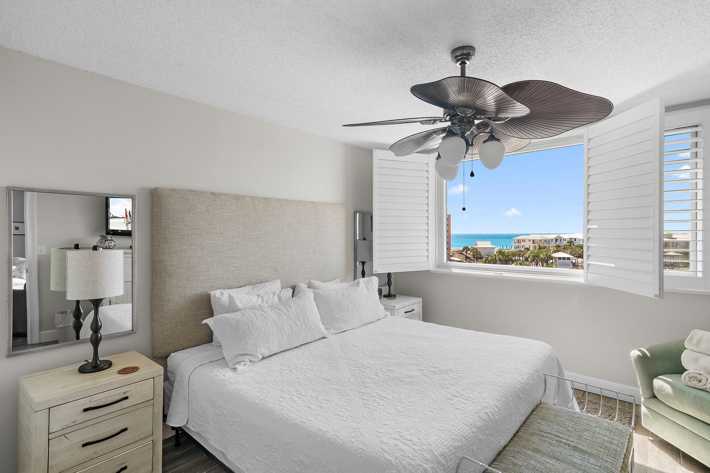 A bedroom with a bed, a nightstand, a lamp, a ceiling fan, a window with blinds, and a view of the ocean.