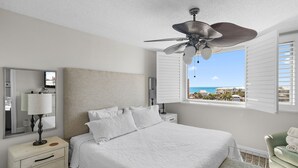 Un dormitorio con cama, mesita de noche, lámpara, ventilador de techo y ventana con persianas, con vista al océano.
