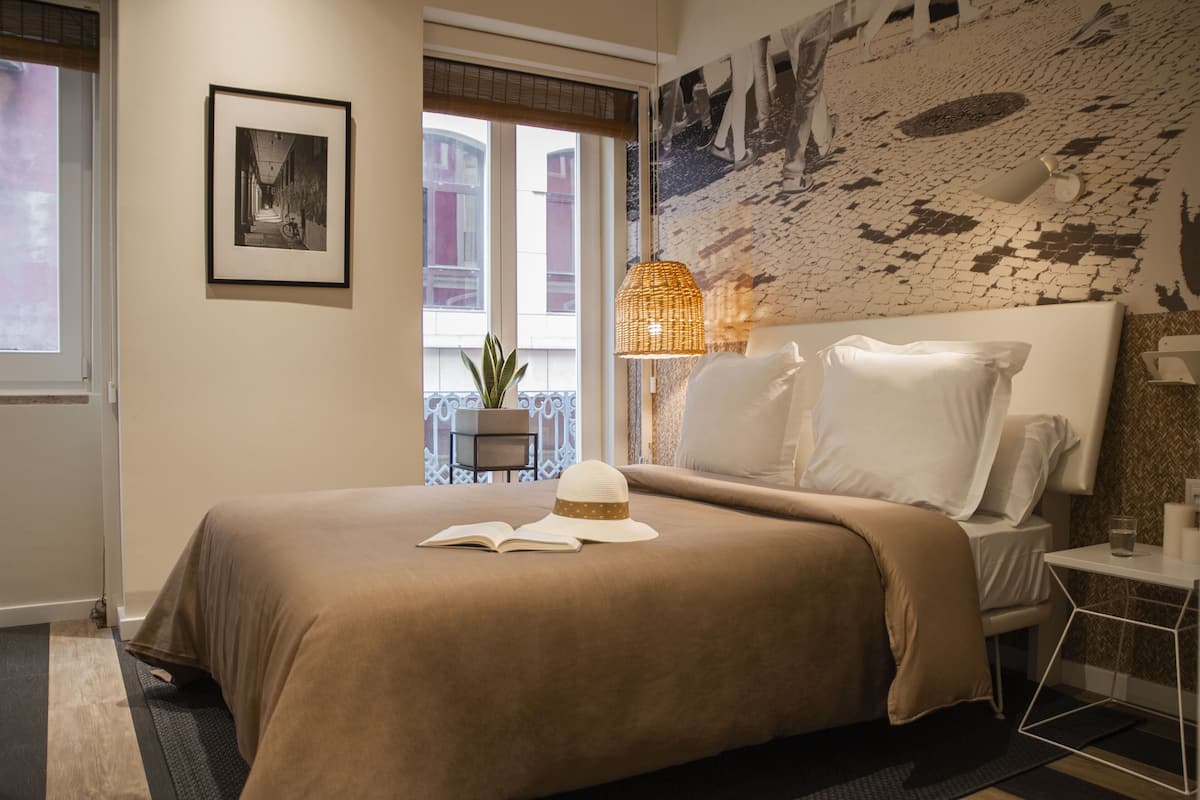 A neatly made bed with a beige blanket and pillows, a small bedside table with a glass of water, a wicker lamp, and a framed picture on the wall.