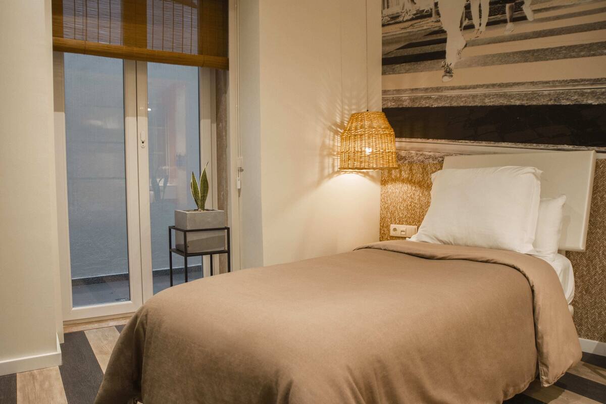 A hotel room with a bed, a nightstand, a lamp, and a view of the outside.