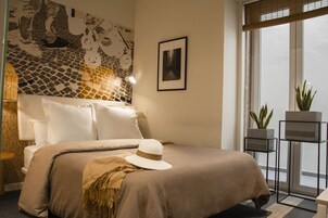 A neatly arranged bedroom with a bed, pillows, a hat, a wall art piece, a framed photo, and two potted plants.