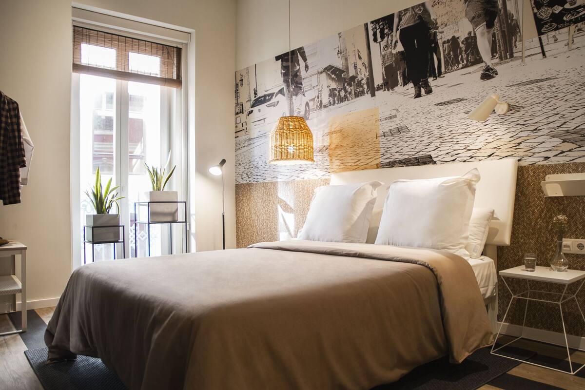 A modern bedroom with a large bed, a cityscape mural on the wall, a window with blinds, and a small side table with a lamp.
