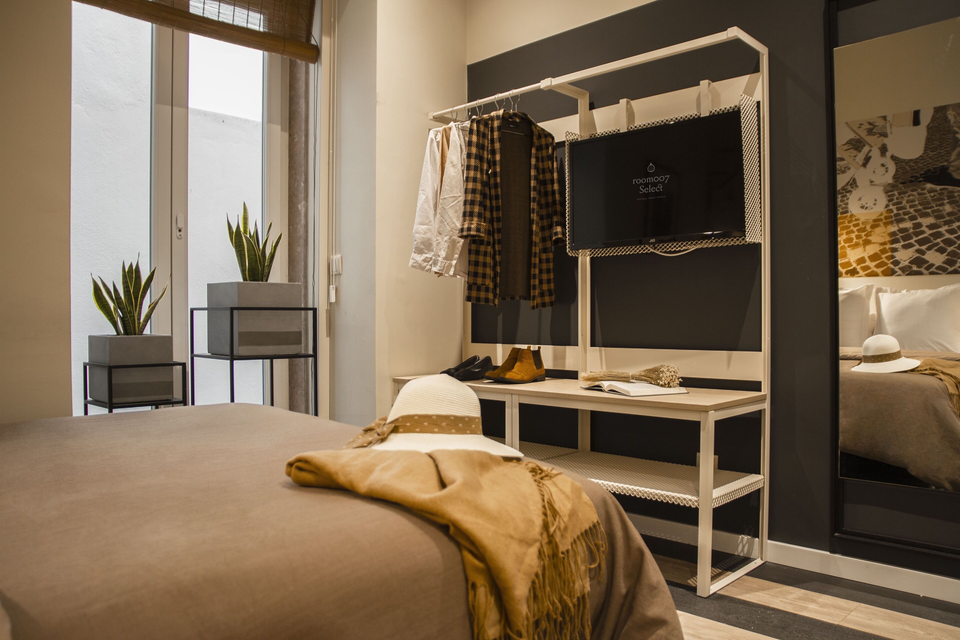 Une chambre d’hôtel moderne avec un grand lit, un bureau avec une chaise, une télévision et une armoire. Des plantes et un miroir ornent les murs.