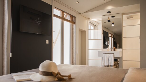 Una camera d'albergo moderna con un letto, un cappello e un libro sul letto. C'è una TV fissata al muro e si intravede un bagno sullo sfondo.