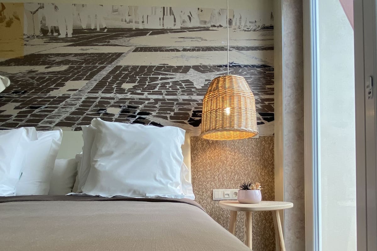 A modern hotel room with a large bed, a hanging light fixture, and a view through a large window.