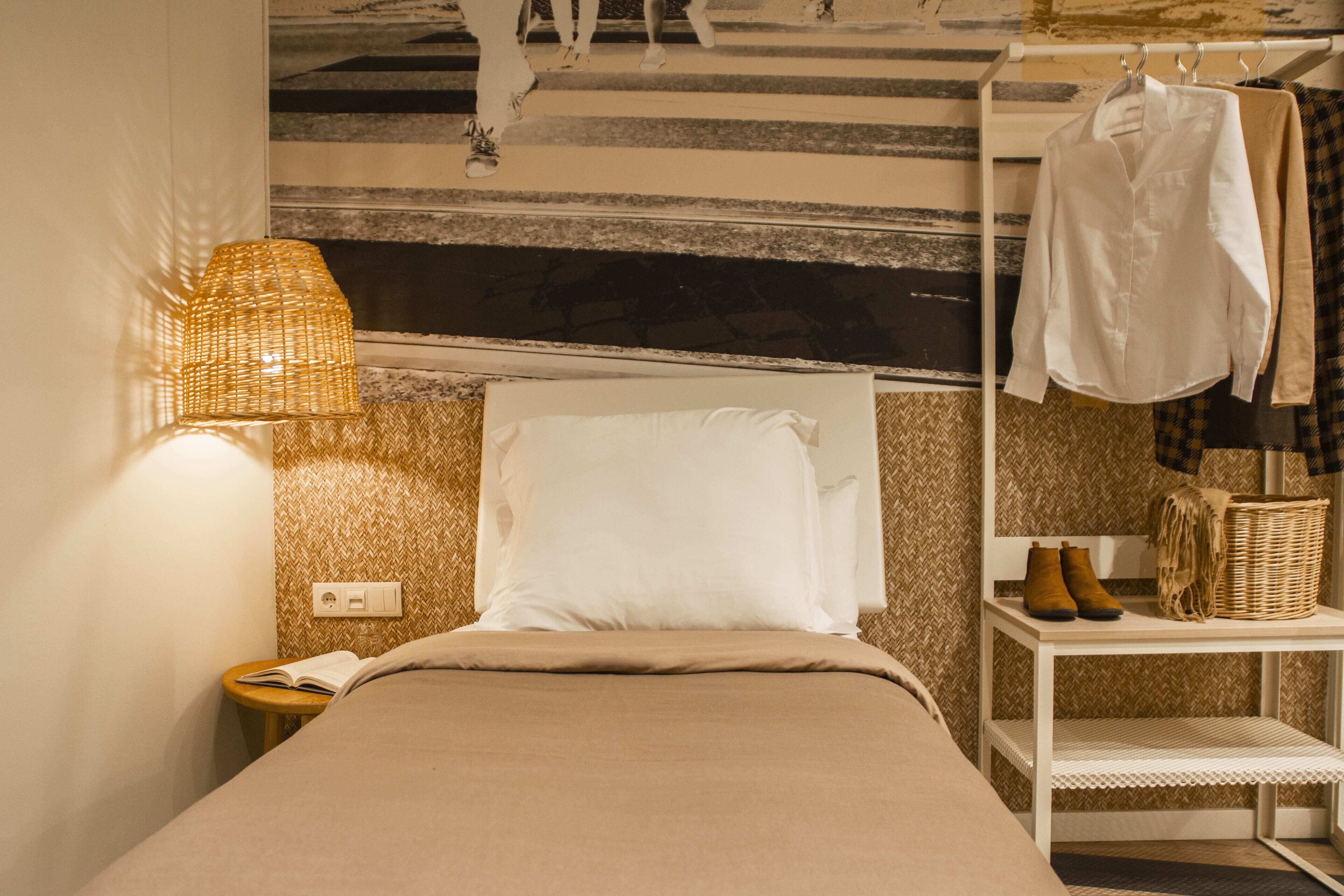 Una cama bien hecha con una almohada blanca y una manta color beige, una lámpara de noche, una pequeña mesita de madera con un libro, y un perchero con una camisa blanca y una camisa de cuadros.