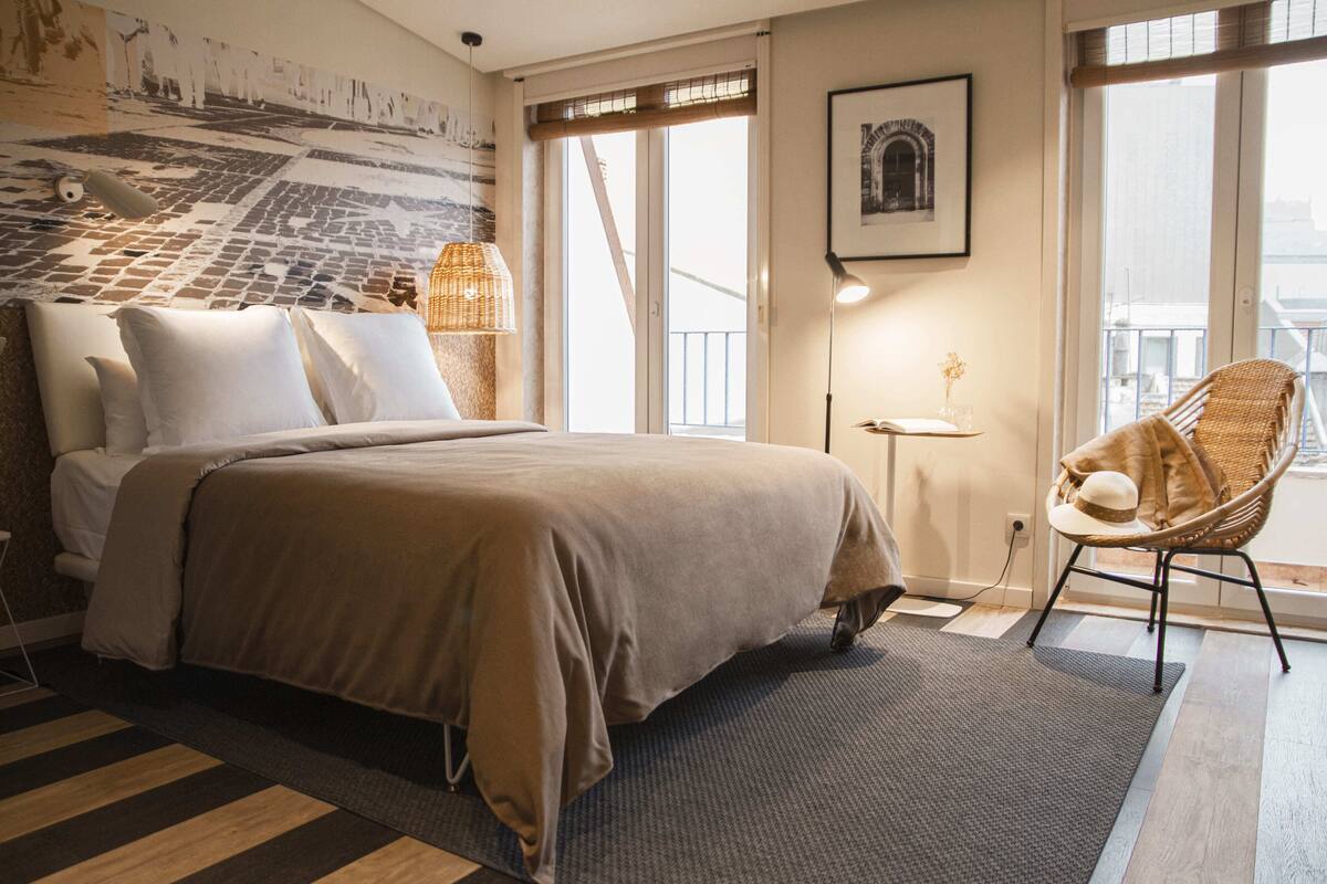 A modern hotel room with a large bed, a wicker chair, a small table, and a view of the city through the window.