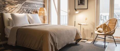 Une chambre d’hôtel moderne avec un grand lit, un fauteuil en osier, une petite table et une vue sur la ville par la fenêtre.