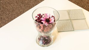 Un vase en verre transparent contenant des fleurs séchées et une seule fleur rose, posé sur une table blanche.