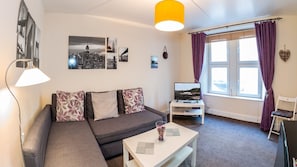 A modern living room with a sectional sofa, a flat-screen TV, and a large window with purple curtains. - Mycosy Lorimer 17 (Dundee)