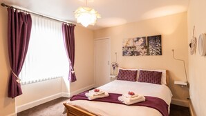 Une chambre à coucher avec un lit, des rideaux violets, une table de chevet avec un vase et une œuvre d’art accrochée au mur.