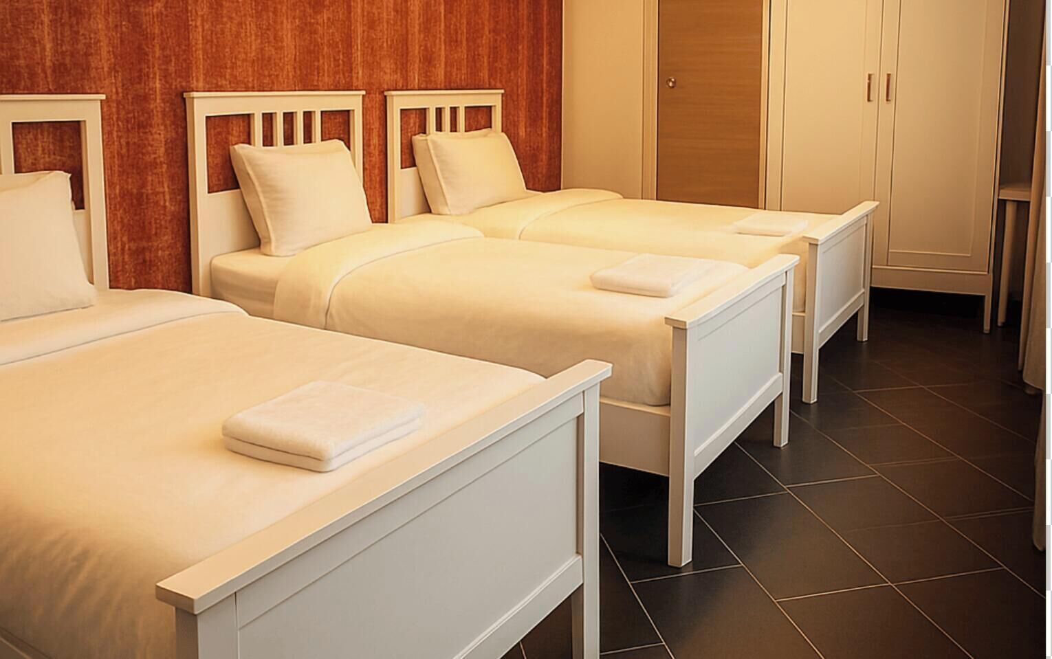 A hotel room with three beds, a wooden wall, a wardrobe, and a tiled floor.