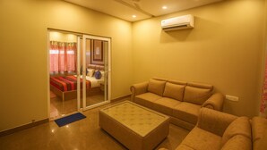 A hotel room with a sofa, a small ottoman, a sliding glass door leading to a bedroom, and an air conditioning unit on the wall. - Virat Resorts, Sariska (Viratnagar)