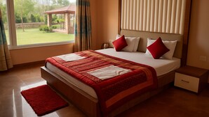 A hotel room with a large bed, a red rug, a wooden nightstand, and a view of a garden and gazebo through the window. - Virat Resorts, Sariska (Viratnagar)