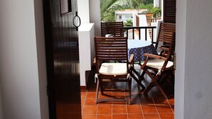 Un patio al aire libre con suelo de baldosas, sillas de madera, una mesa y vistas a un edificio y palmeras.