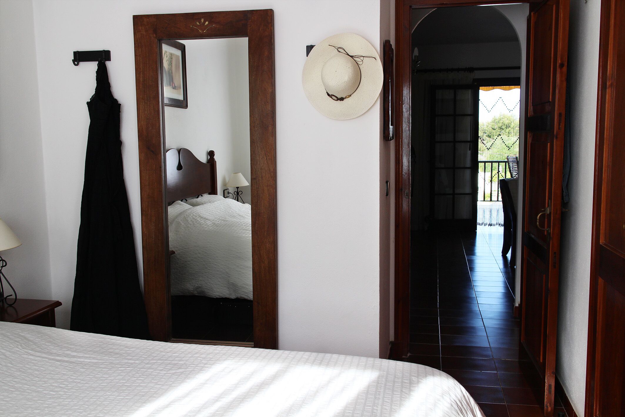 A bedroom with a bed, a hanging black garment, a mirror, and a hat on the wall.