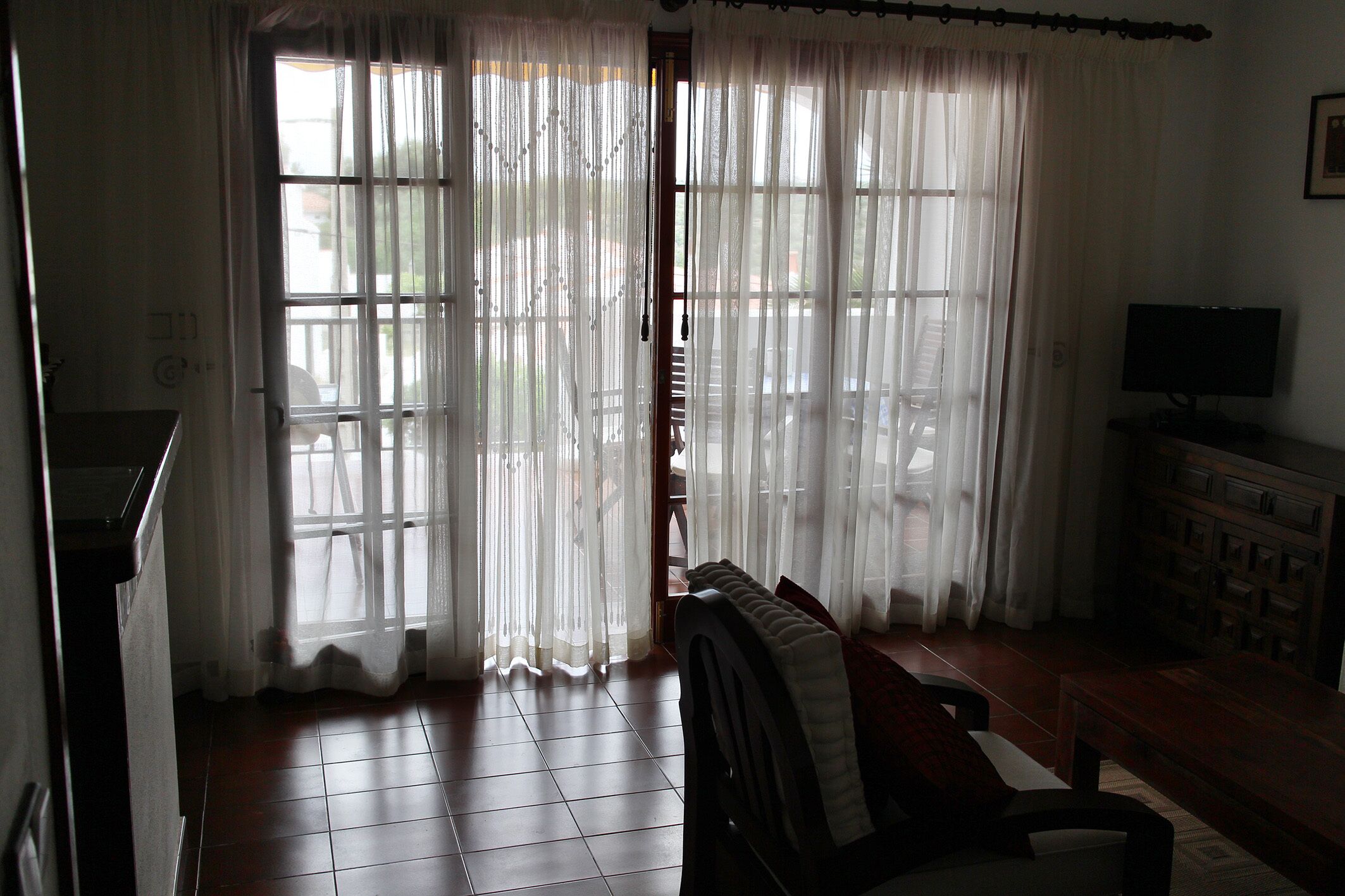A room with a view through sheer curtains, a television, and a tiled floor.