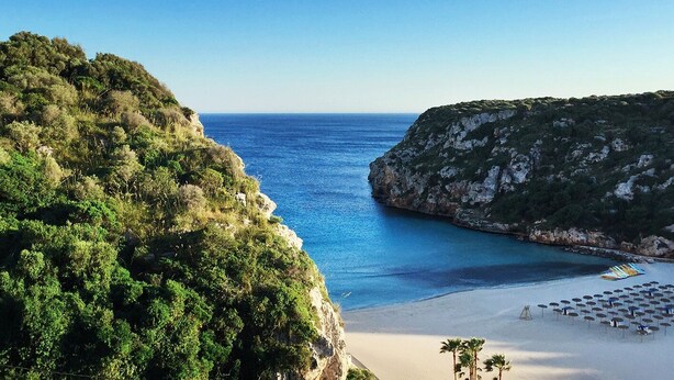 Una playa costera con un acantilado rocoso y una zona de arena con sombrillas y tumbonas.