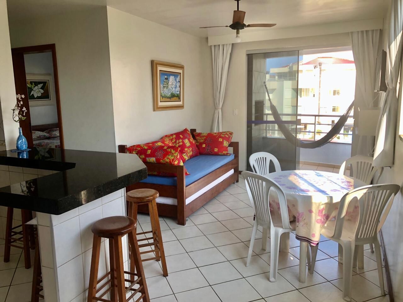 Un espacio habitable compacto con kitchenette, un sofá con cojines rojos, una mesa de comedor con sillas y un ventanal con cortinas.