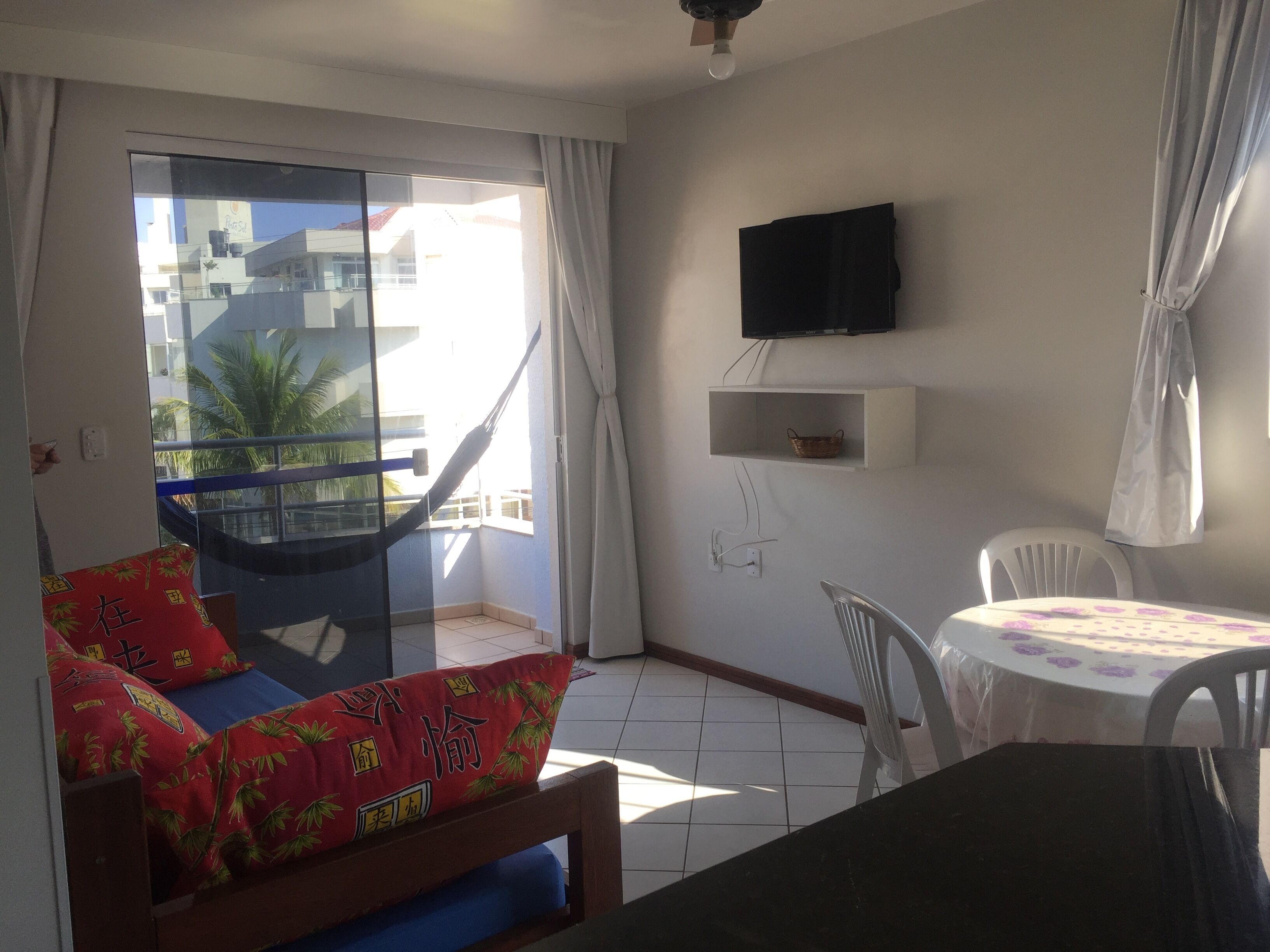 A balcony with a hammock, a small table with chairs, a TV on the wall, and a window with curtains.