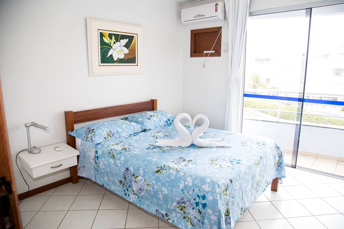 A bedroom with a bed, a nightstand, a wall-mounted air conditioner, a window with a view, and a framed picture on the wall.