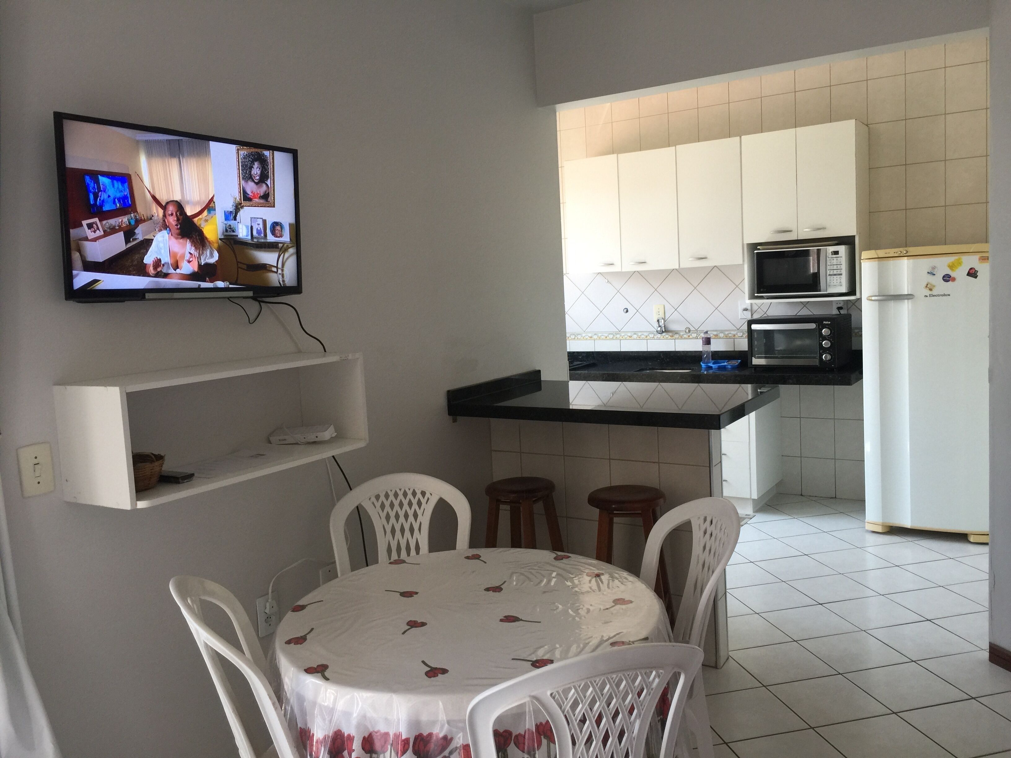 Eine kompakte Küche mit weißen Schränken, einer Mikrowelle und einem Kühlschrank. Es gibt einen Esstisch mit Stühlen und einen an der Wand montierten Fernseher.