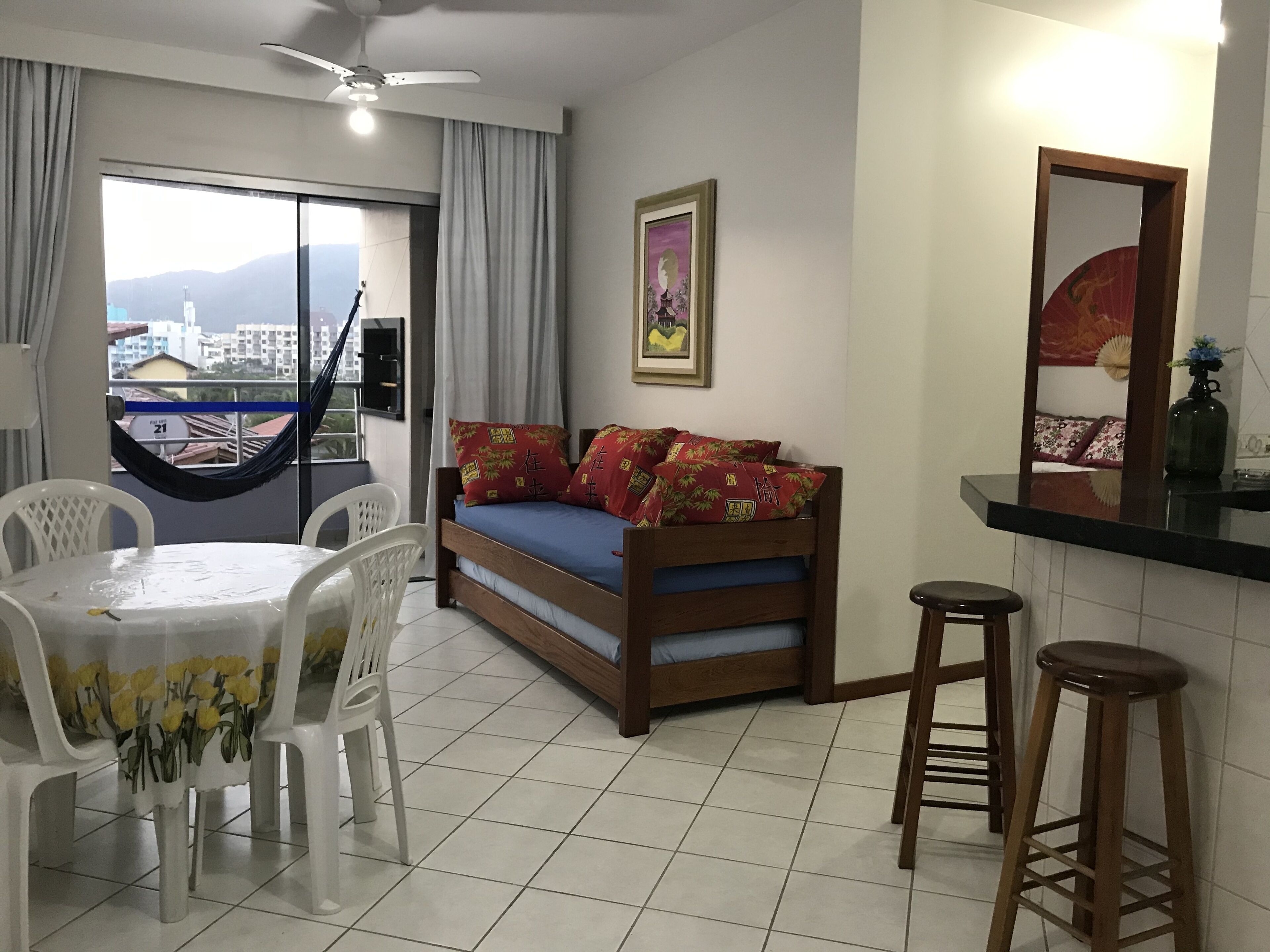 Um quarto com uma cama, uma mesa de jantar com cadeiras, uma varanda com vista para edifícios e um ventilador de teto.