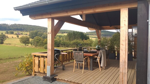Una terraza de madera con zona de estar cubierta, una parrilla y vistas a un paisaje verde.