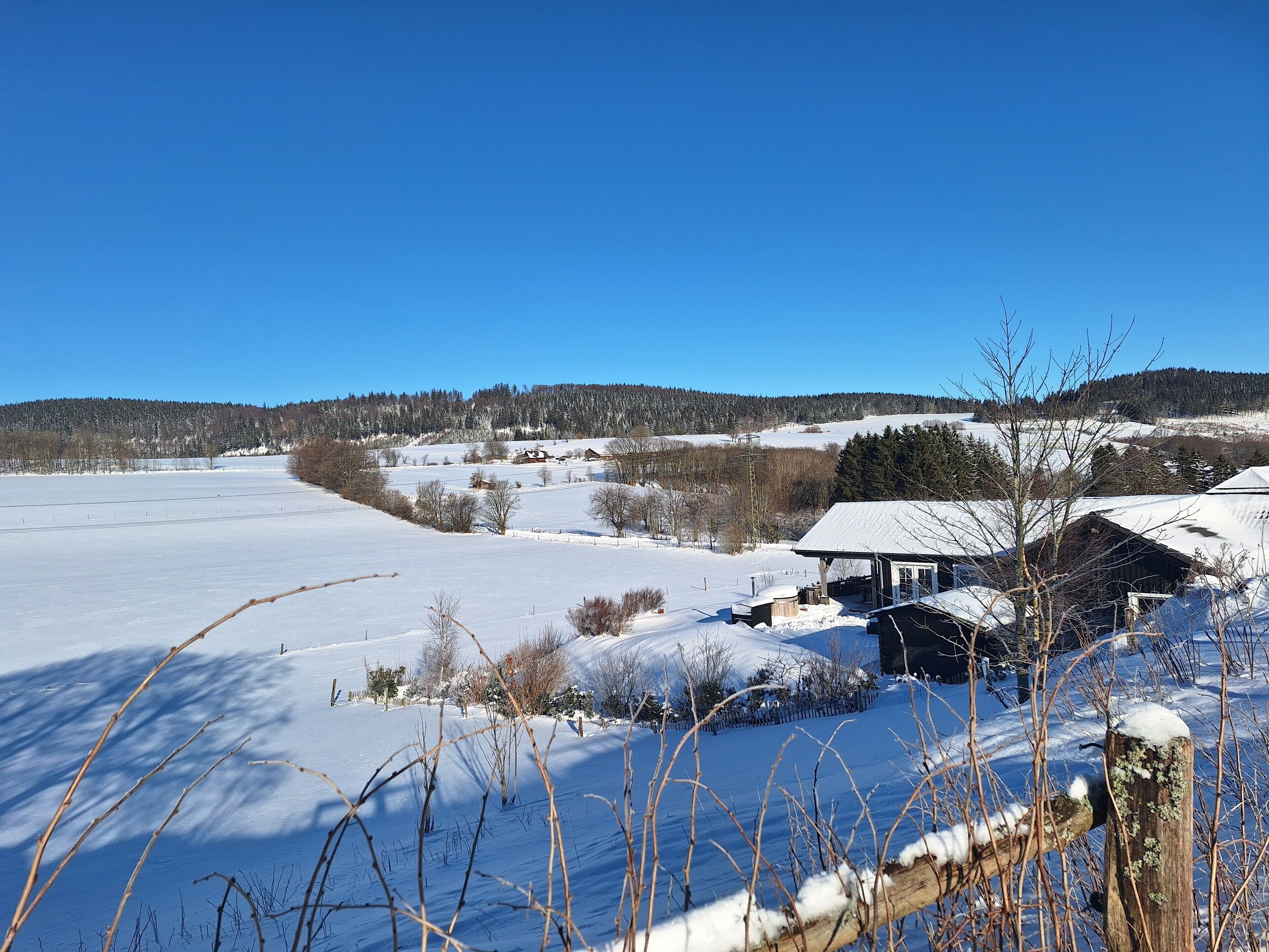 A rural winter scene with a snow-covered field, a wooden cabin, and a clear blue sky.