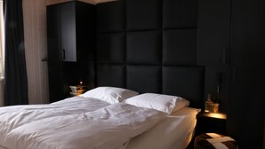 Un dormitorio con una cama, cabecera oscura, mesita de noche con lámpara y un reposapiés pequeño y redondo.