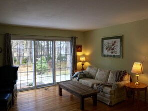 A living room with a sofa, a coffee table, and a lamp. There is a window with curtains, a framed picture on the wall, and a view of trees outside.