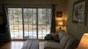 Sala de estar com mesa de madeira, sofá, abajur e vista para uma área arborizada por grandes janelas.