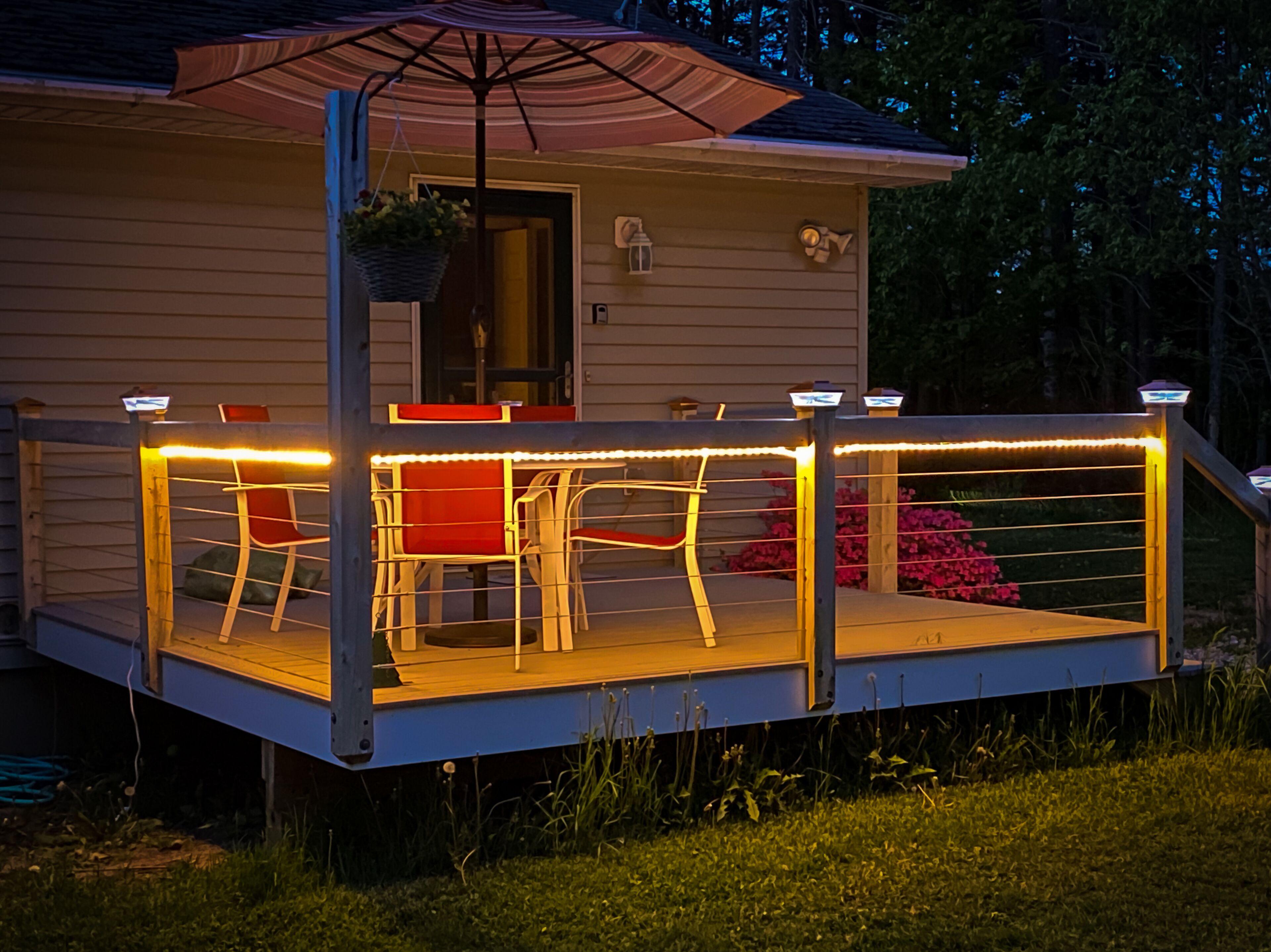 Une terrasse extérieure couverte, dotée d’une table et de chaises, d’un pot de fleurs suspendu et d’un petit espace porche avec une porte et des fenêtres.