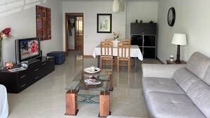 Een moderne woonkamer met een bank, een glazen salontafel, een televisie en een eethoek met een tafel en stoelen.